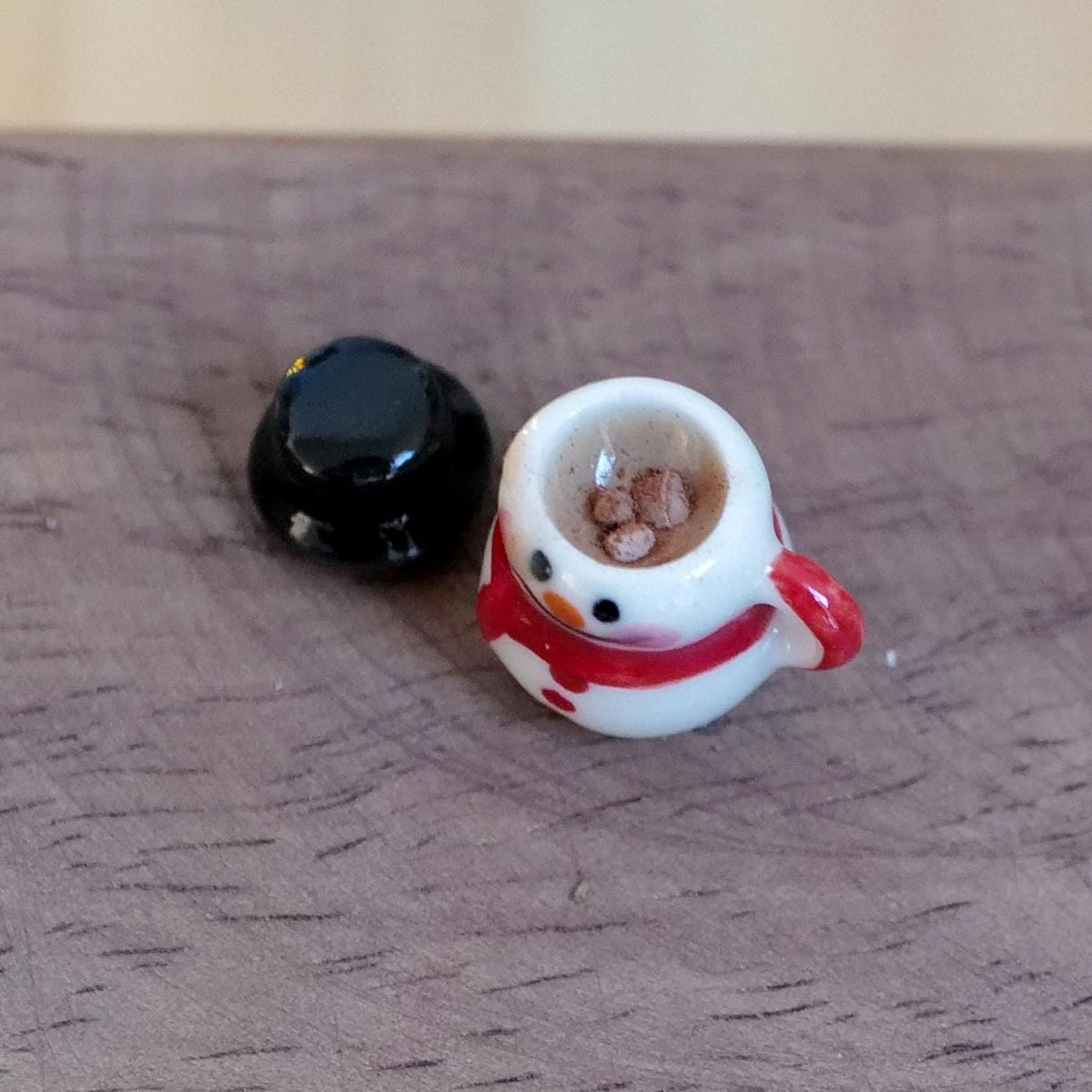1:12 scale miniature white snowman mug with a red scarf, filled with hot chocolate and marshmallows, with its black top hat lid beside it on a wood table
