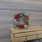 1:12 scale miniature red rose and berry wreath with cream rosebuds and sage sprigs on a twig base, hanging loop on top, shown on a wood wall