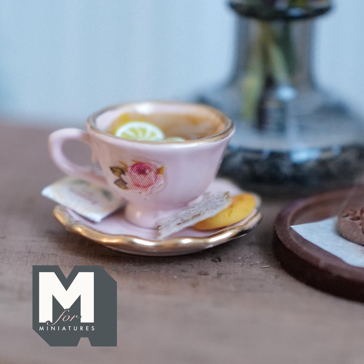 1:12 scale miniature pink cup and saucer with gold rims and a pink rose, filled with lemon tea and lemon slices, with a tea bag and a jam cookie on the saucer