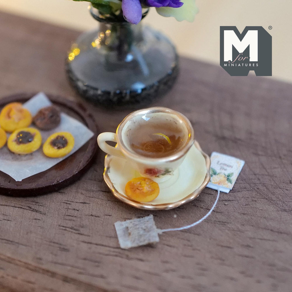 1:12 scale miniature cream cup and saucer with gold rims and a pink rose, filled with tea, with a jam cookie on the saucer and a tea bag on a string beside it