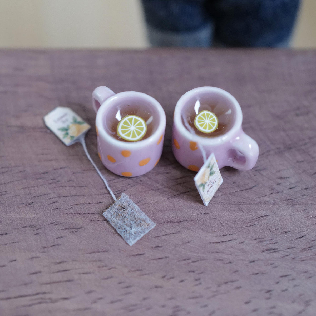 Two 1:12 scale miniature pink mugs with orange polka dots, filled with tea and lemon slices, with tea bags beside them on a wood table
