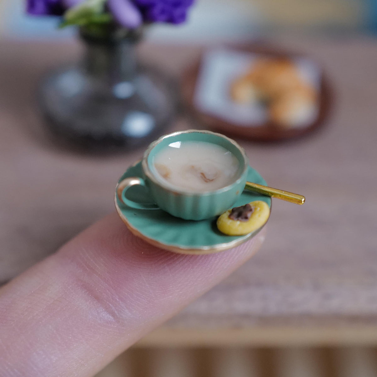 1:12 scale miniature green fluted coffee cup with gold rim, filled with milky stirred coffee and a gold-tone spoon, on a matching saucer with a jam thumbprint cookie, held on a fingertip
