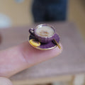 1:12 scale miniature purple fluted coffee cup with gold rim, filled with milky stirred coffee and a gold-tone spoon, on a matching saucer with a jam thumbprint cookie, held on a fingertip