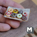 1:12 scale miniature tray of eight iced ring donuts in chocolate, pink, white with sprinkles, yellow and green on parchment on an aged metal baking tray