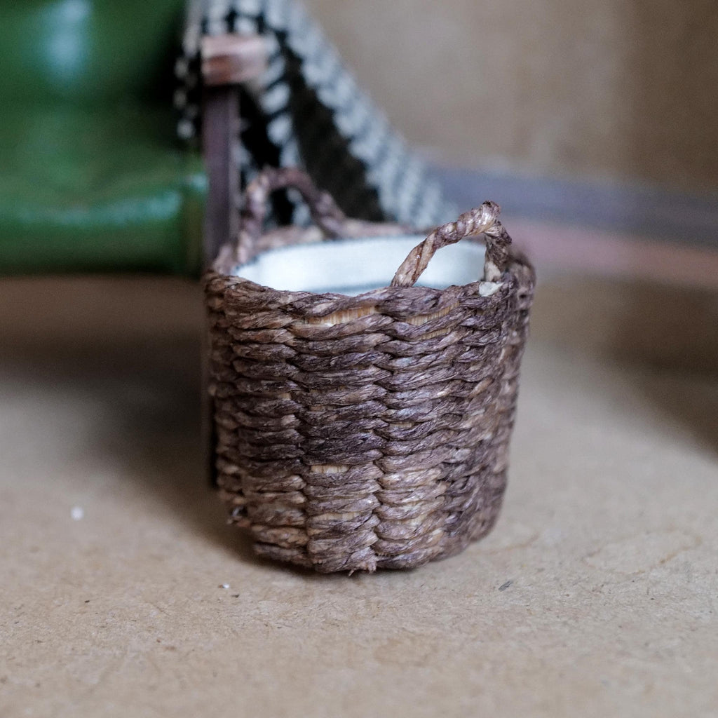 1:12 scale miniature deep woven blanket basket in weathered dark brown with a white linen lining and loop handles, beside a green armchair