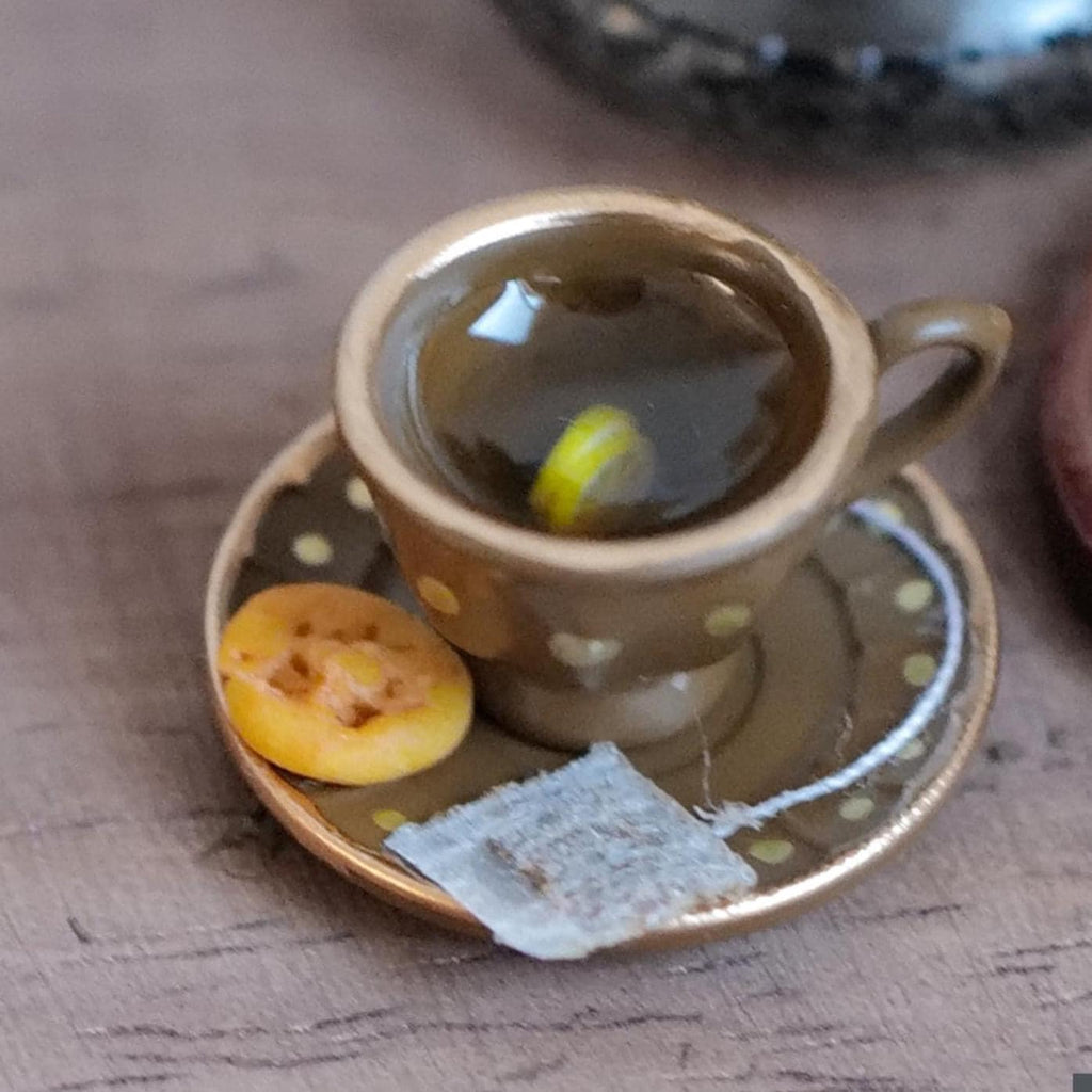 1:12 scale miniature brown tea cup and saucer with gold polka dots and gold rims, filled with lemon tea and a lemon slice, with a tea bag and waffle cookie