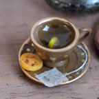 1:12 scale miniature brown tea cup and saucer with gold polka dots and gold rims, filled with lemon tea and a lemon slice, with a tea bag and waffle cookie