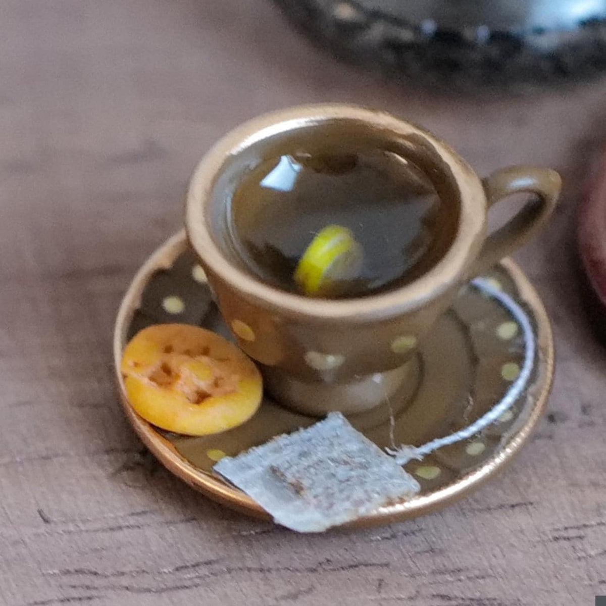 1:12 scale miniature brown tea cup and saucer with gold polka dots and gold rims, filled with lemon tea and a lemon slice, with a tea bag and waffle cookie