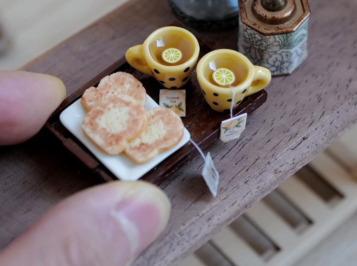1:12 scale miniature wood tea tray with two yellow mugs with black polka dots filled with lemon tea, and sugar cookies on a white plate, held on a fingertip