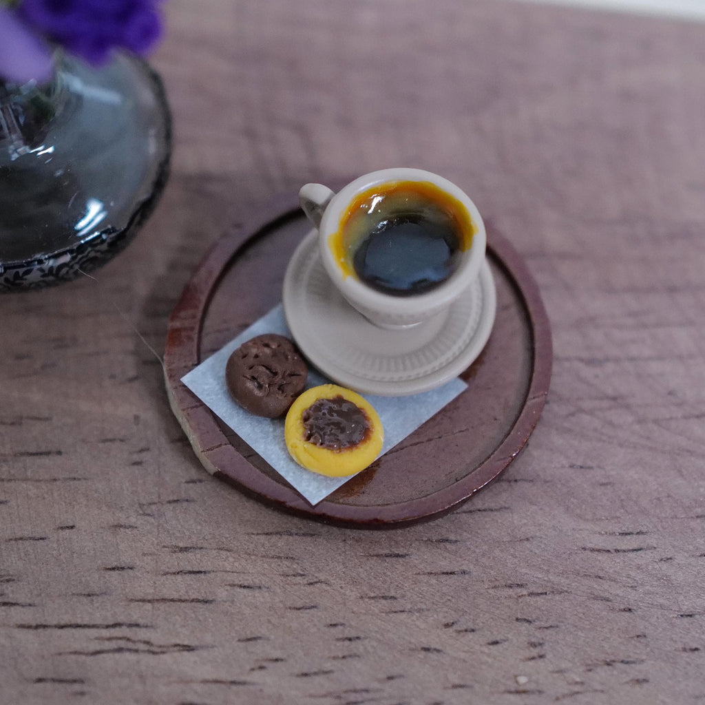 1:12 scale miniature beige cup of black coffee with crema on a saucer, with two chocolate cookies on deli paper, on a round brown serving tray