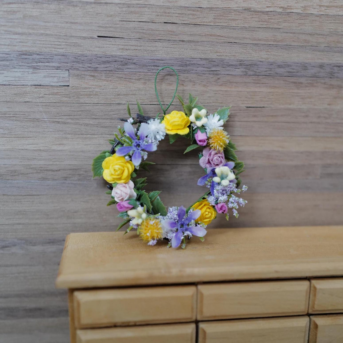 1:12 scale miniature spring flower wreath with yellow and pink roses, purple blooms and lilac sprigs on a twig base, hanging loop on top, shown on a wood wall