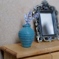 1:12 scale miniature turquoise ribbed clay vase with white and lilac flower stems, beside an ornate mirror on a wood side table