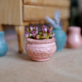 1:12 scale miniature clay potted plant, a round pink pot with an embossed leaf band holding pink and green flower buds, on a wood floor