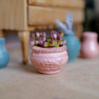 1:12 scale miniature clay potted plant, a round pink pot with an embossed leaf band holding pink and green flower buds, on a wood floor