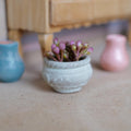 1:12 scale miniature clay potted plant, a weathered light green round pot holding pink and green flower buds, on a wood floor