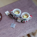 Two 1:12 scale miniature white mugs with a red cherry pattern and red handles, filled with tea and lemon slices, with tea bags beside them on a wood table