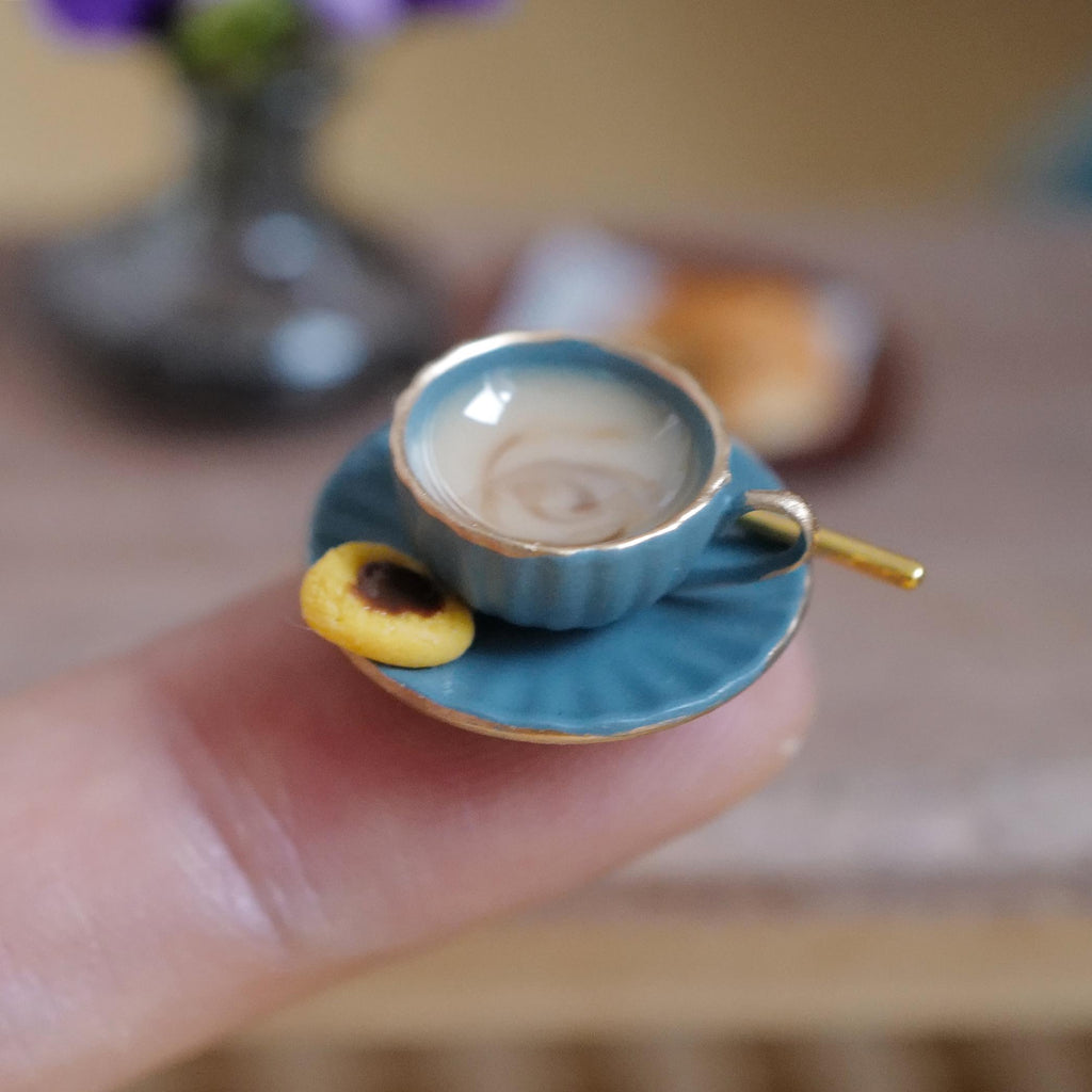 1:12 scale miniature blue fluted coffee cup with gold rim, filled with milky stirred coffee and a gold-tone spoon, on a matching saucer with a jam thumbprint cookie, held on a fingertip