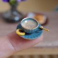 1:12 scale miniature blue fluted coffee cup with gold rim, filled with milky stirred coffee and a gold-tone spoon, on a matching saucer with a jam thumbprint cookie, held on a fingertip