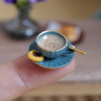 1:12 scale miniature blue fluted coffee cup with gold rim, filled with milky stirred coffee and a gold-tone spoon, on a matching saucer with a jam thumbprint cookie, held on a fingertip