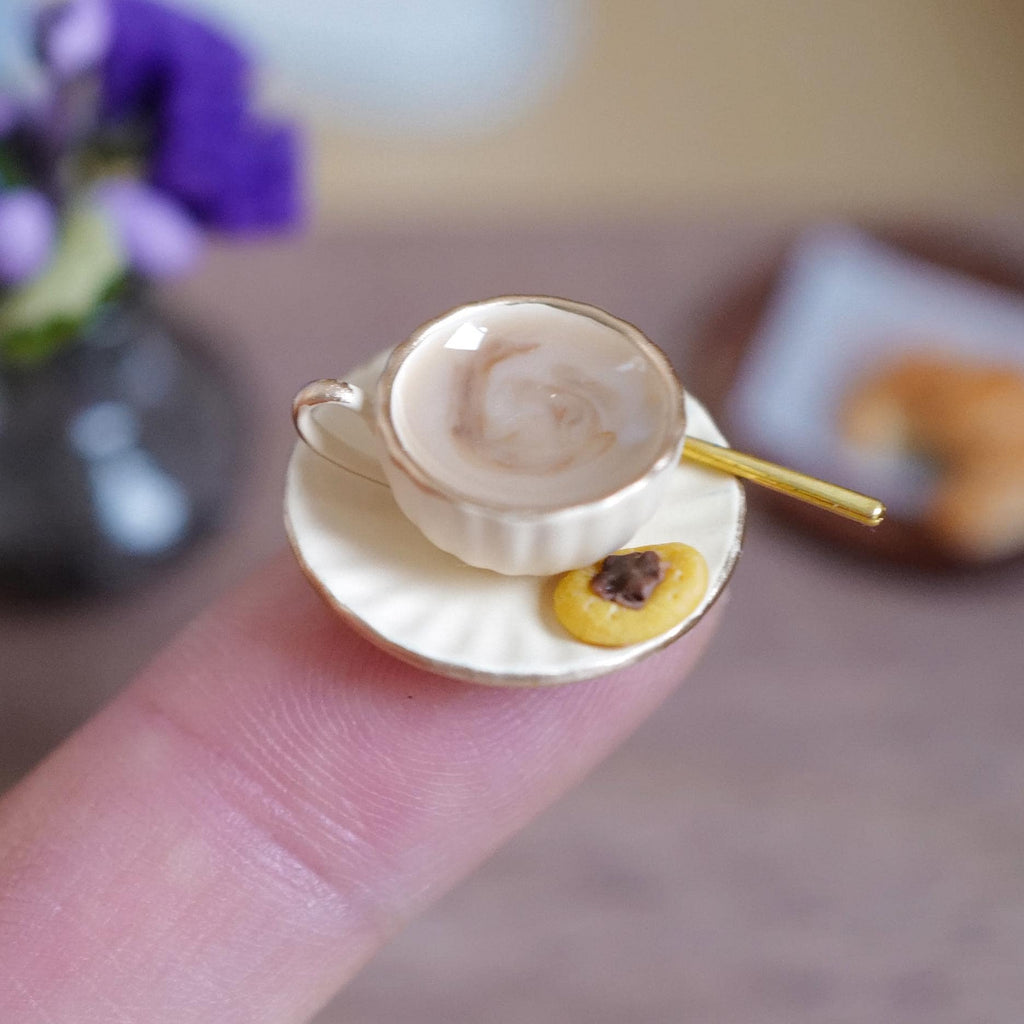 1:12 scale miniature cream fluted coffee cup with gold rim, filled with milky stirred coffee and a gold-tone spoon, on a matching saucer with a jam thumbprint cookie, held on a fingertip