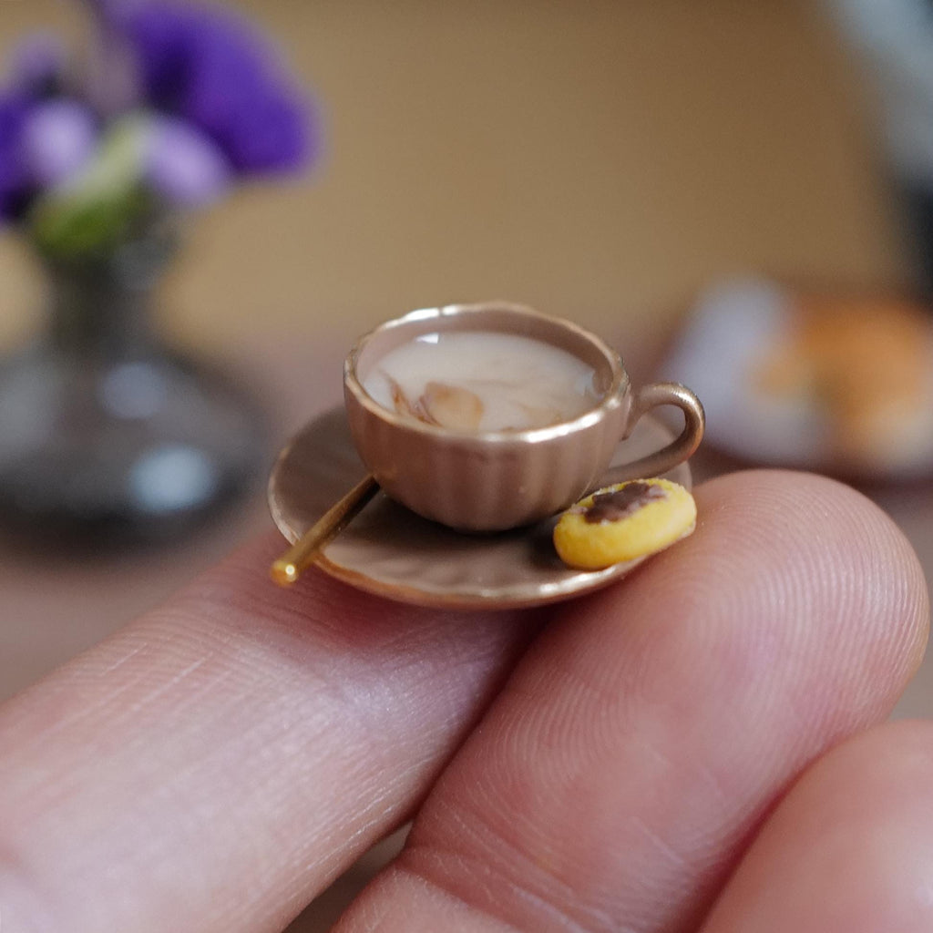 1:12 scale miniature brown fluted coffee cup with gold rim, filled with milky stirred coffee and a gold-tone spoon, on a matching saucer with a jam thumbprint cookie, held on a fingertip