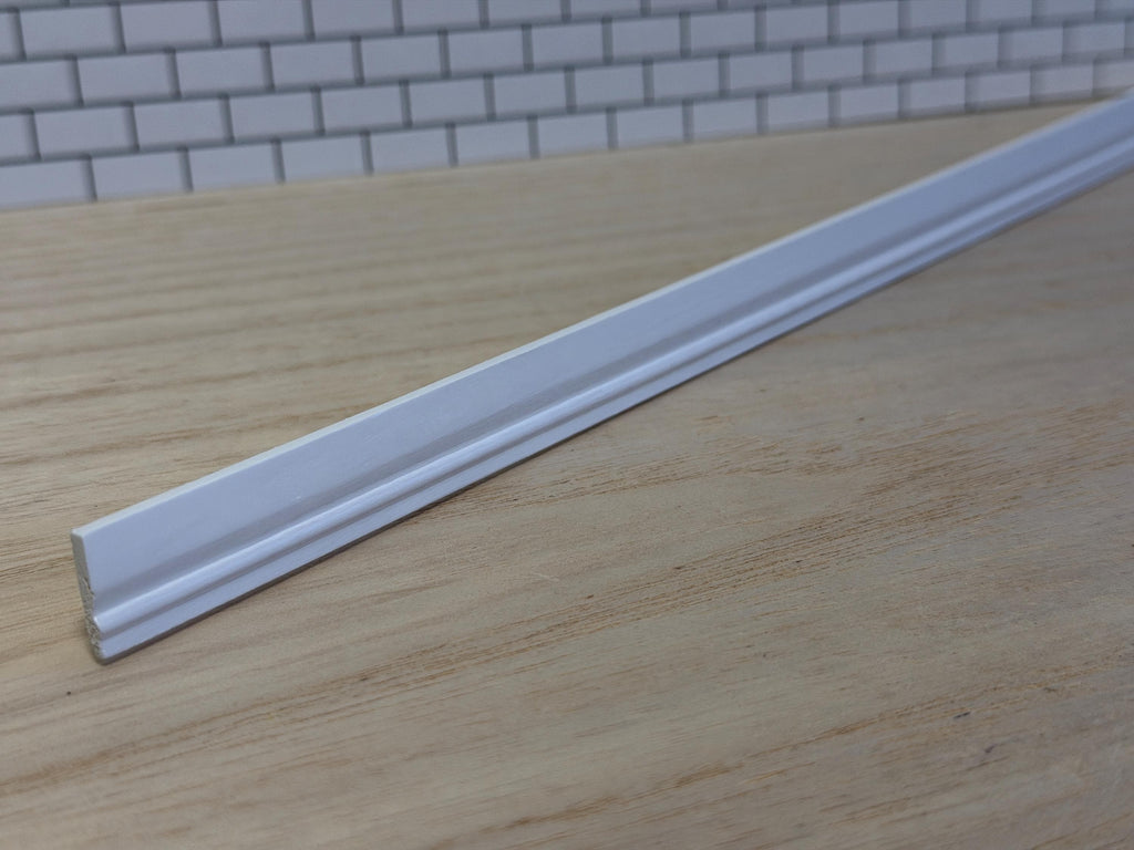 1:12 scale miniature white wood baseboard molding lying on a wood surface in front of a white brick wall, showing its curved profile and stepped lower edge along the full length