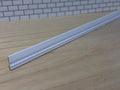 1:12 scale miniature white wood baseboard molding lying on a wood surface in front of a white brick wall, showing its curved profile and stepped lower edge along the full length