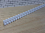 1:12 scale miniature white wood baseboard molding lying on a wood surface in front of a white brick wall, showing its curved profile and stepped lower edge along the full length