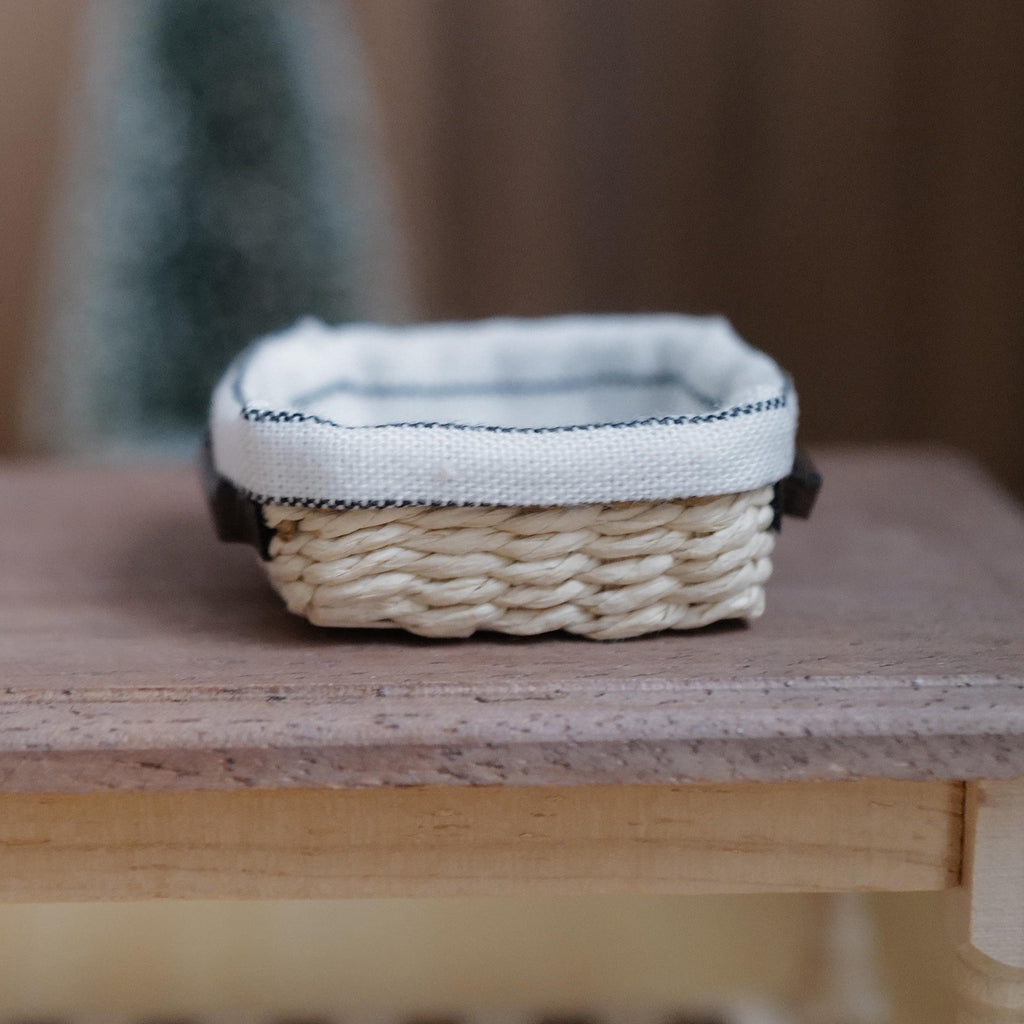 1:12 scale miniature shallow rectangular woven bread basket with a white striped linen lining and dark brown faux leather handles on a wood table